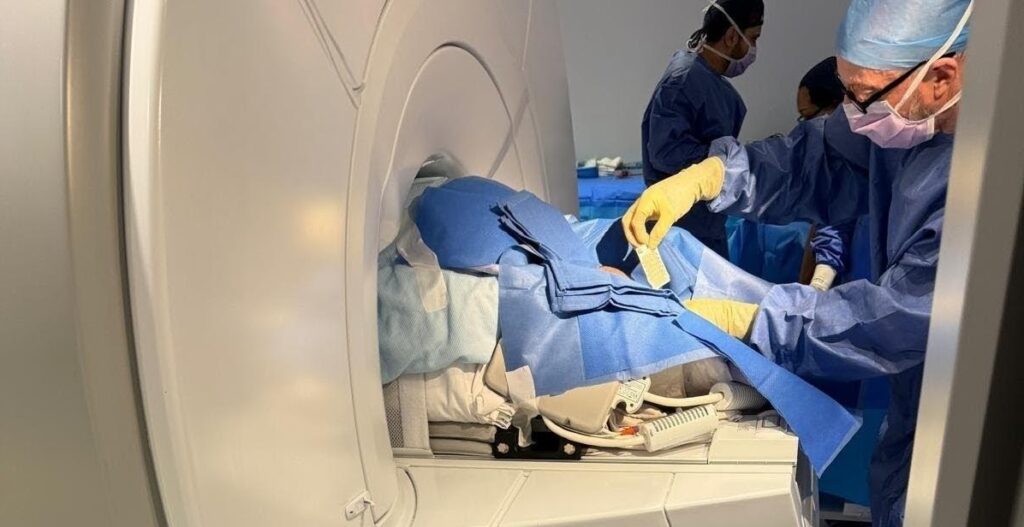 Mae'r ddelwedd yn dangos claf yng nghanol llawdriniaeth mewn sganiwr MRI gyda'r niwrolawfeddyg mewn cap, mwgwd, gŵn a menig yn eu paratoi ar gyfer llawdriniaeth.